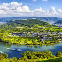 A wide shot of a bend in the Moselle River from it's surrounding hills under broad daylight, two river cruise ships can be seen creating gentle waves in the water.