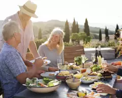 A group of mature friends are sitting around an outdoor dining table in Tuscany, Italy